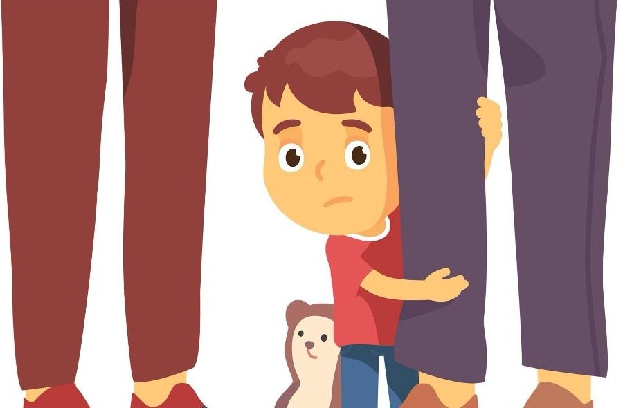 A young child hiding behind a parent’s legs, holding a soft toy